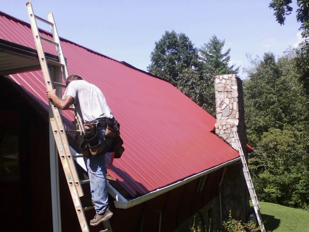 Roofer on a ladder beside a red metal roof and stone chimney — Soffit & Fascia Repair by Roofing Brothers in Orem